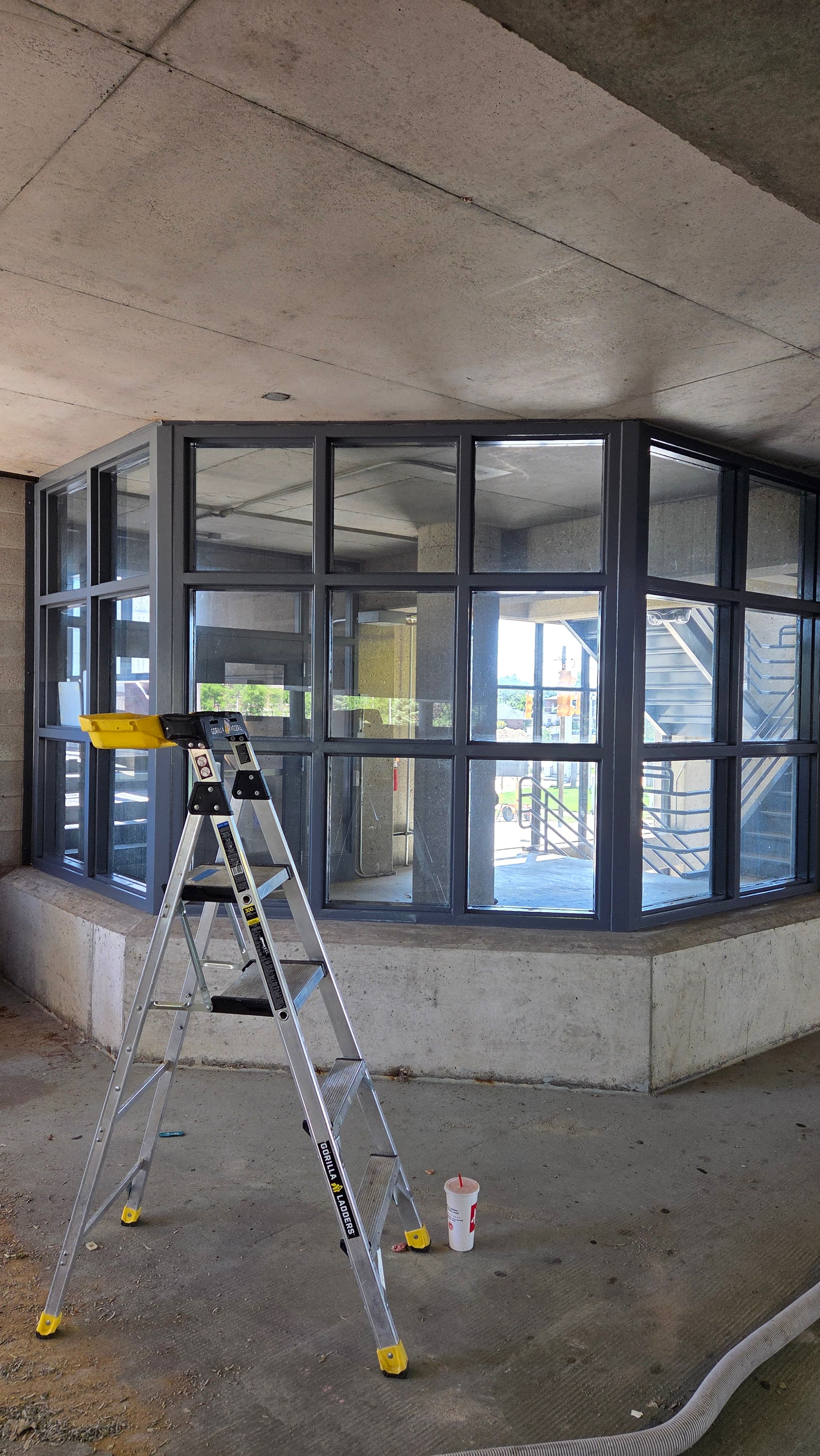 Silver step ladder in front of a large, dark-framed window under a concrete ceiling.